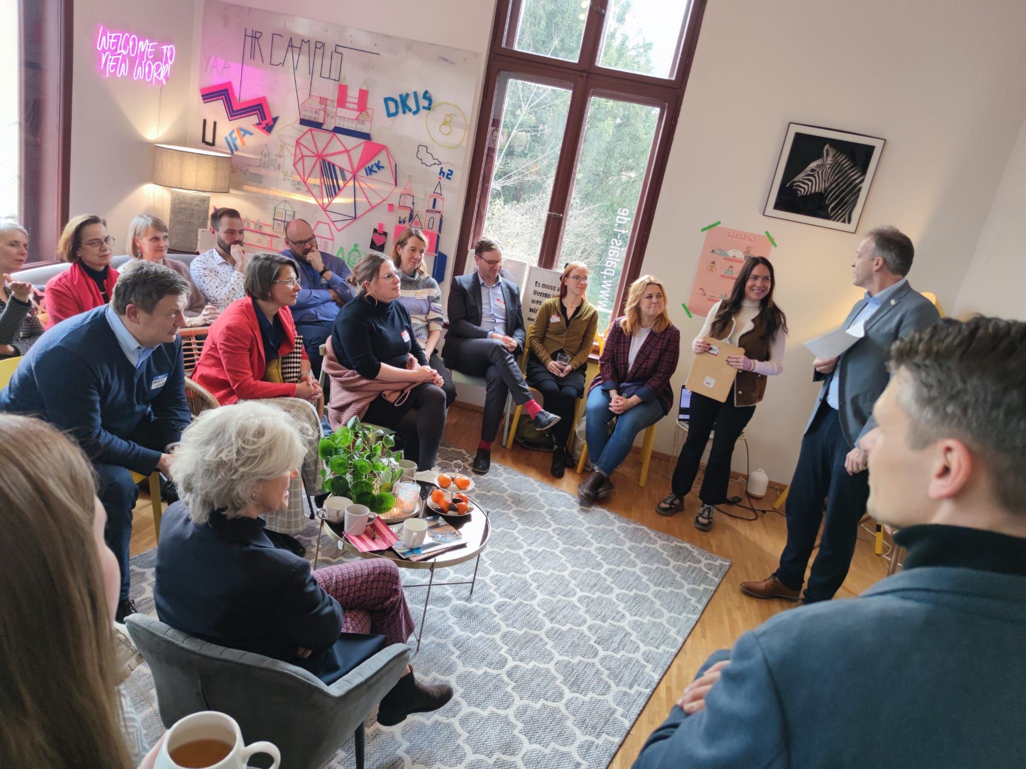 Menschen sitzen und stehen in einem Raum. Alle schauen zu einer Person rechts im Bild, die gerade redet