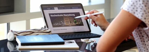 Auf einem Schreibtisch liegen Bücher und ein Tablet mit der Hochschule Merseburg auf dem Display. Ein Stift zeigt auf das Display.