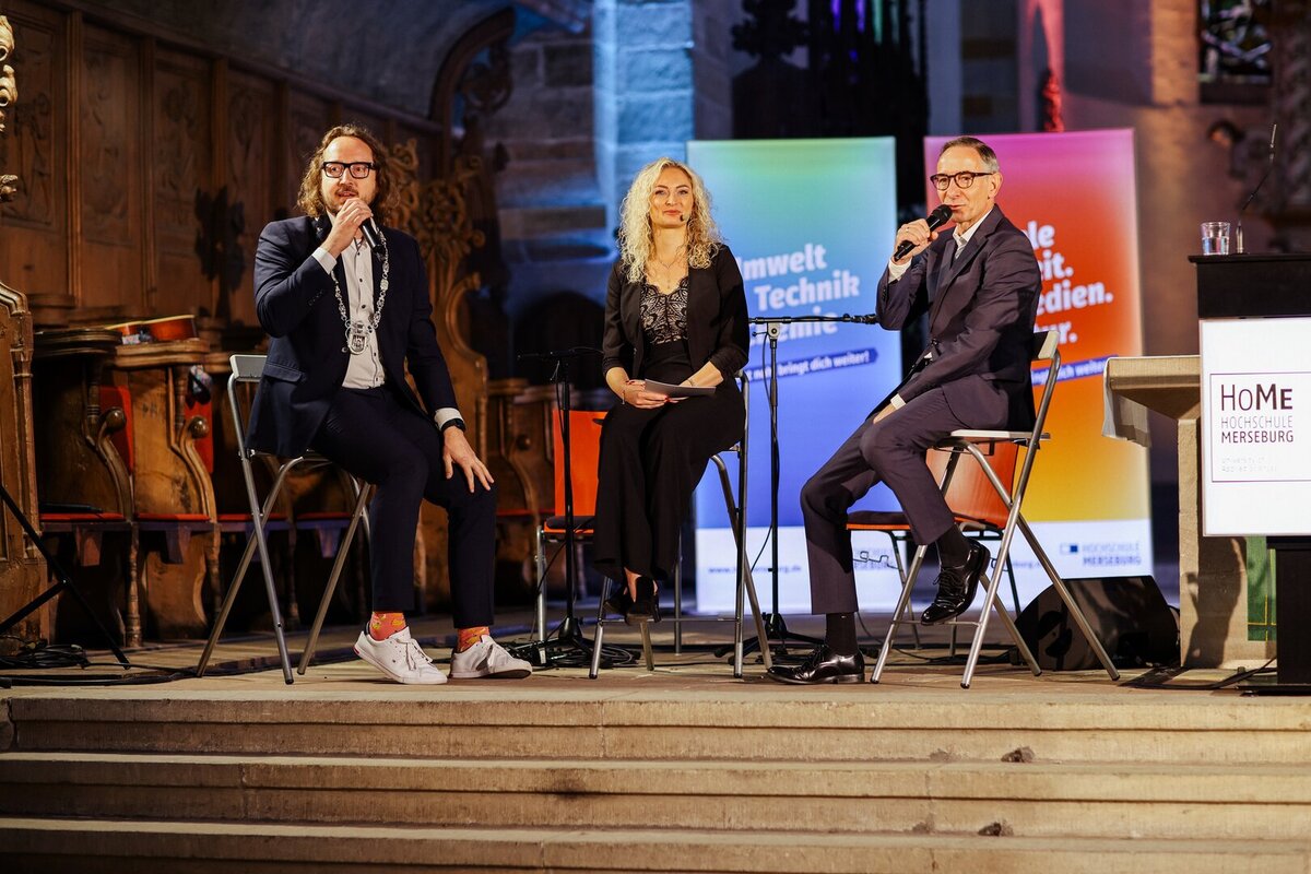 3 Personen auf Barhockern am ALtar mit buntem Hintergrund
