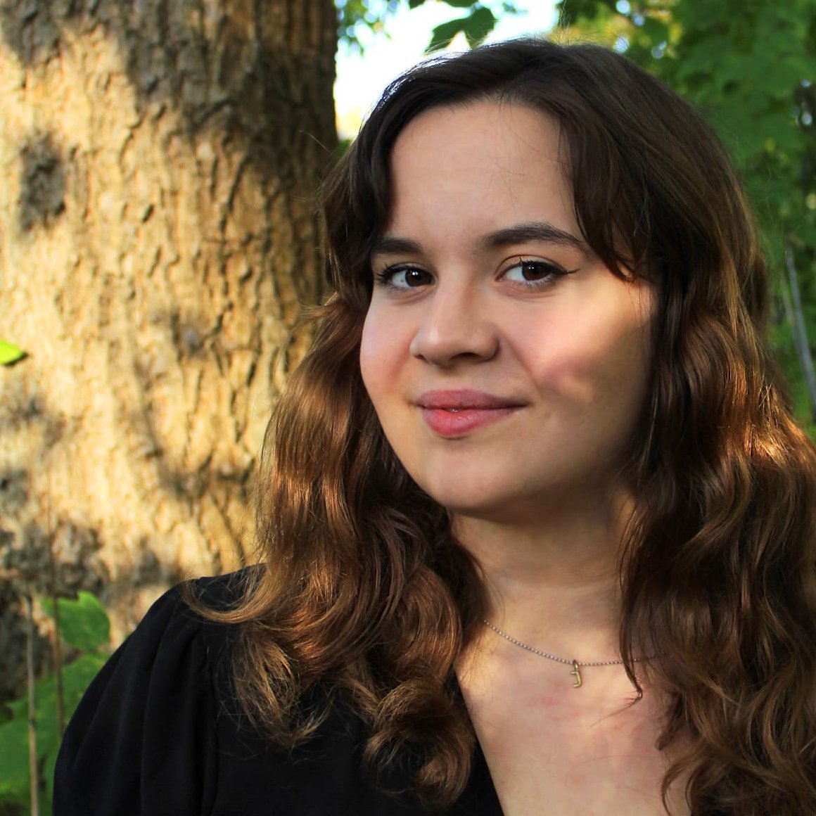 Eine Frau steht vor einem Baum. Sie hat braune Harre, lächelt und hat ihren Kopf ist leicht im Profil geneigt. Die Frau ist bis zur Brust zu sehen