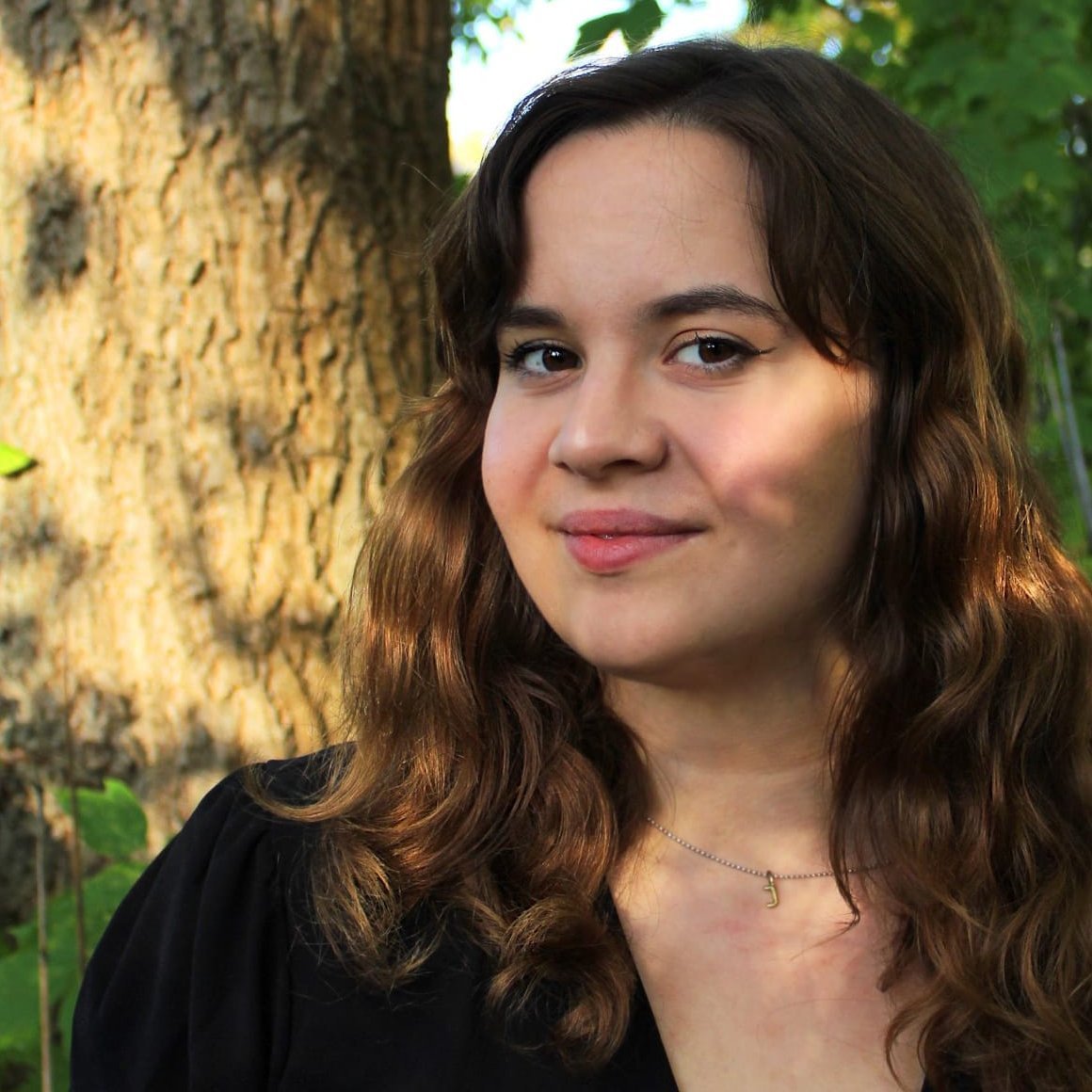 Eine Frau steht vor einem Baum. Sie hat braune Harre, lächelt und hat ihren Kopf ist leicht im Profil geneigt. Die Frau ist bis zur Brust zu sehen