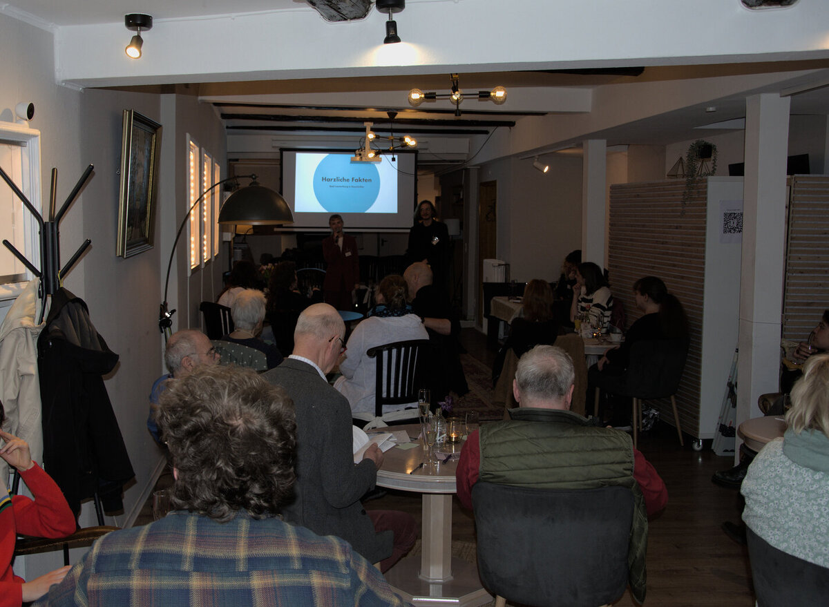 Blick in ein kleines und dunkles Café mit heller Leinwand im Hintergrund zentral im Bild. Auf die Leinwand schauen Menschen verschiedenem Alters. Sie sitzen in Gruppen an runden Tischen