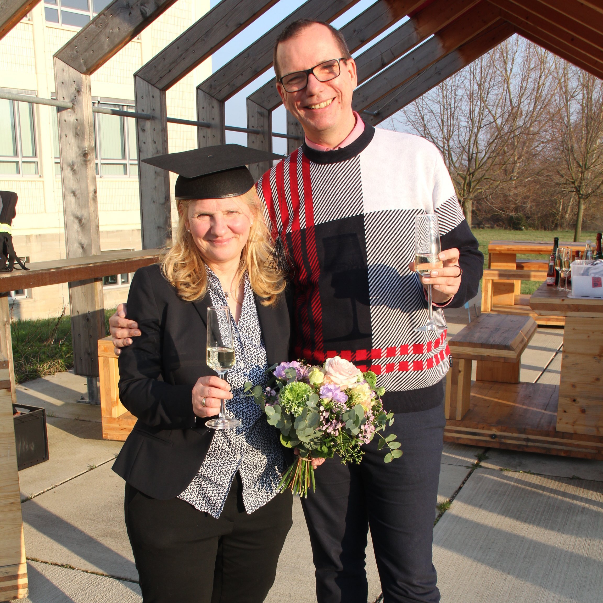 Auf dem Bild sind zwei Personen zu sehen. Links eine Frau mit Blumenstrauß und Doktorhut, rechts ein Mann mit einem Sektglas in der Hand