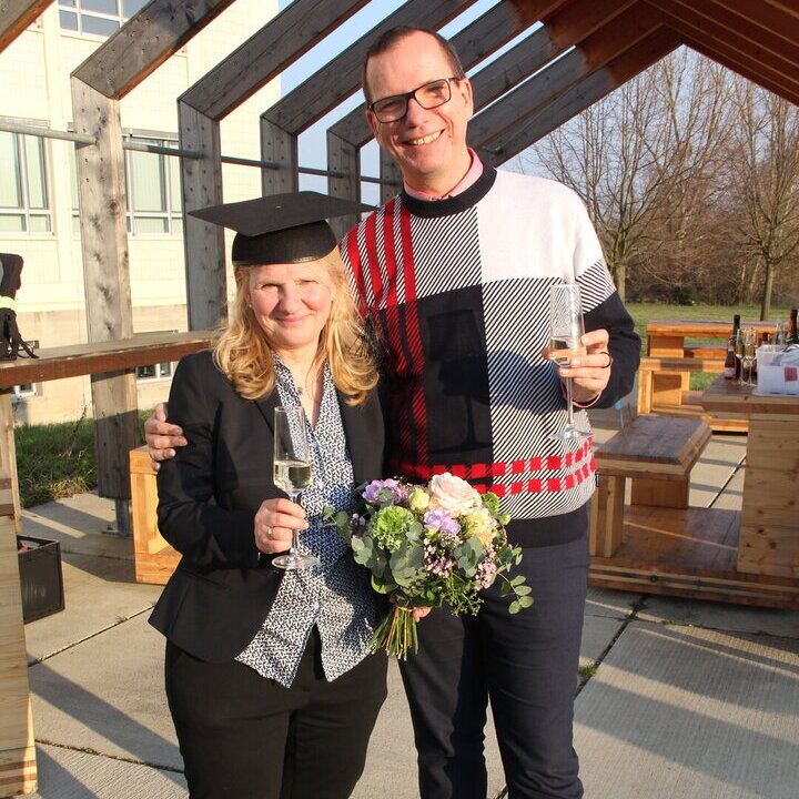 Auf dem Bild sind zwei Personen zu sehen. Links eine Frau mit Blumenstrauß und Doktorhut, rechts ein Mann mit einem Sektglas in der Hand