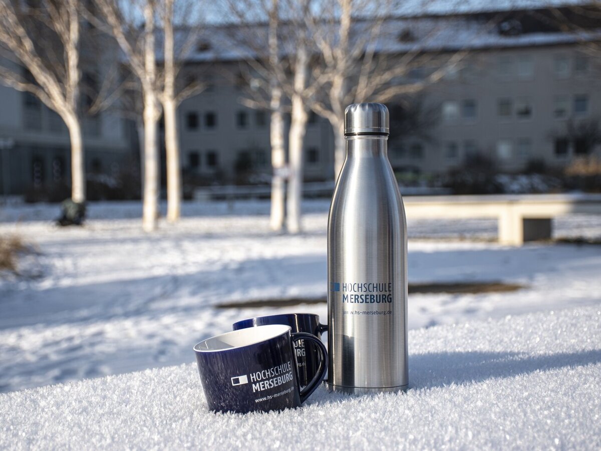 Eine blaue Tasse neben einer silbernen Isolierflasche mit Hochschullogo im Schnee