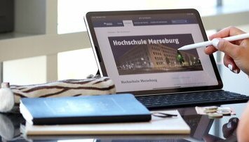 Auf einem Schreibtisch liegen Bücher und ein Tablet mit der Hochschule Merseburg auf dem Display. Ein Stift zeigt auf das Display.