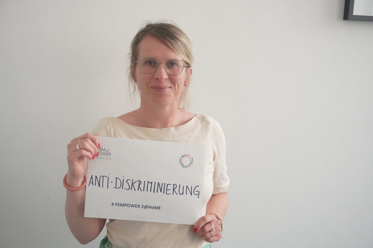 Eine weiblich gelesene Person steht vor einer weißen Wand und trägt ein Schild mit dem Aufdruck Anti-Diskriminierung 