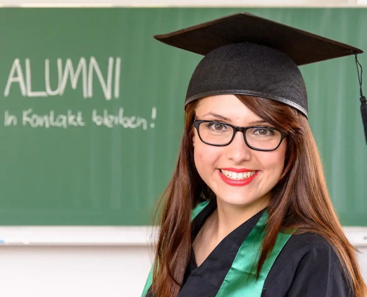 Eine Frau im Talar steht vor einer Tafel mit Aufschrift: Alumni - in Kotakt bleiben!