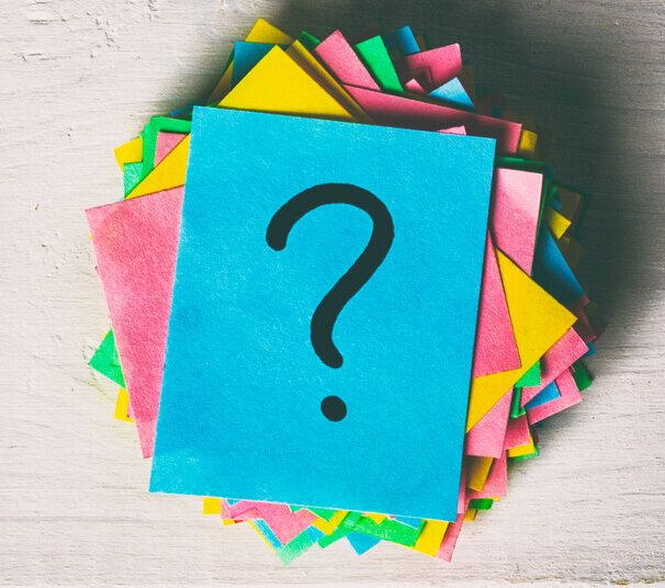 Ein Stapel bunter Noitzzettel liegt rechts im Bild auf Holzleisten. Auf dem obersten Zettel ist ein Fragezeichen zu sehen