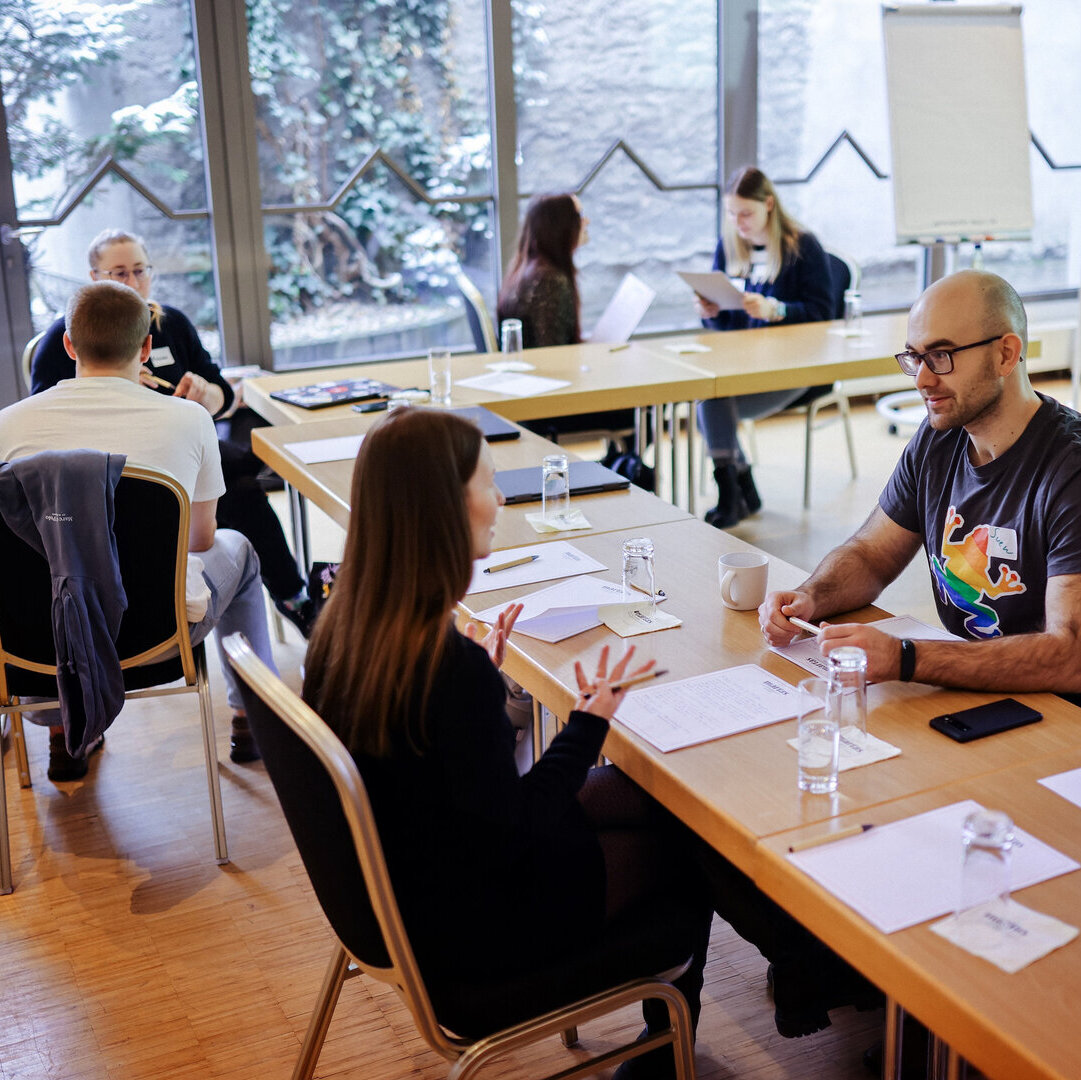 In einem Seminarraum stehen mehrere Tische in L-Form. An den Tischen sitzen Menschen in Zweier-Gruppen und unterhalten sich.