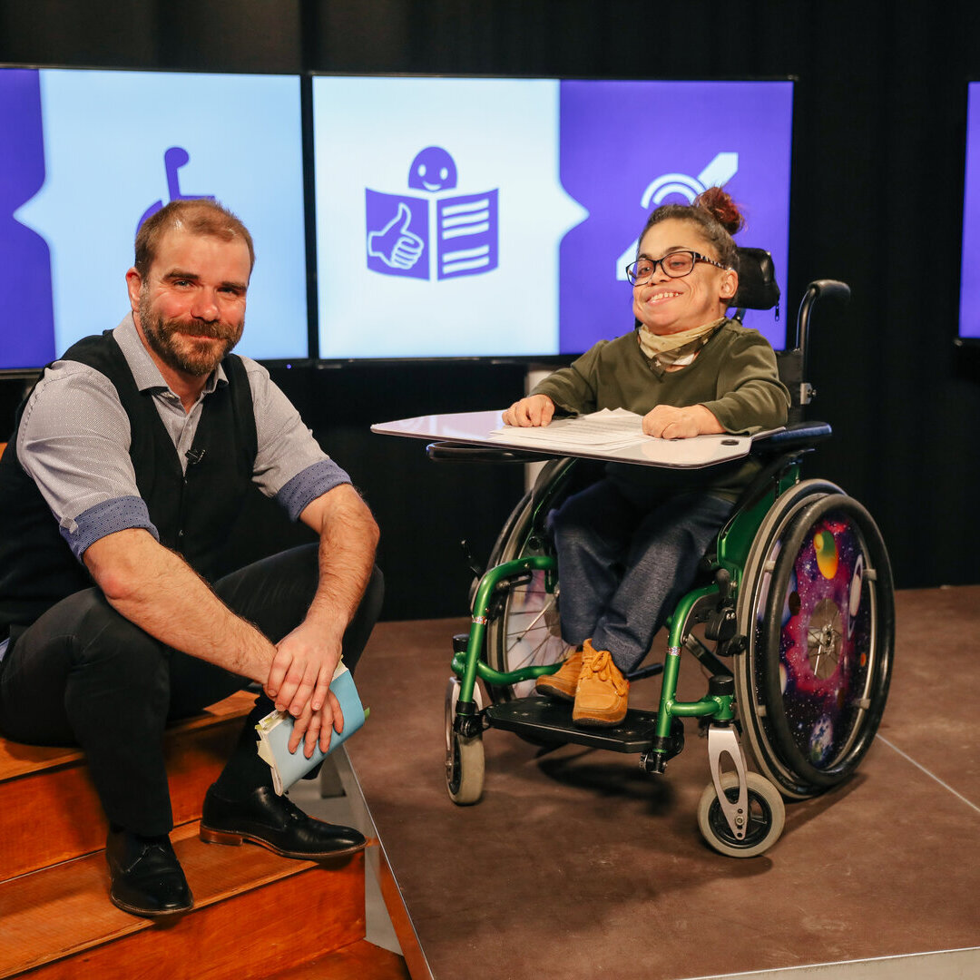 Das Bild zeigt eine Person mit einer körperlichen Behinderung im Rollstuhl neben einer sitzenden Person vor Bildschirmen