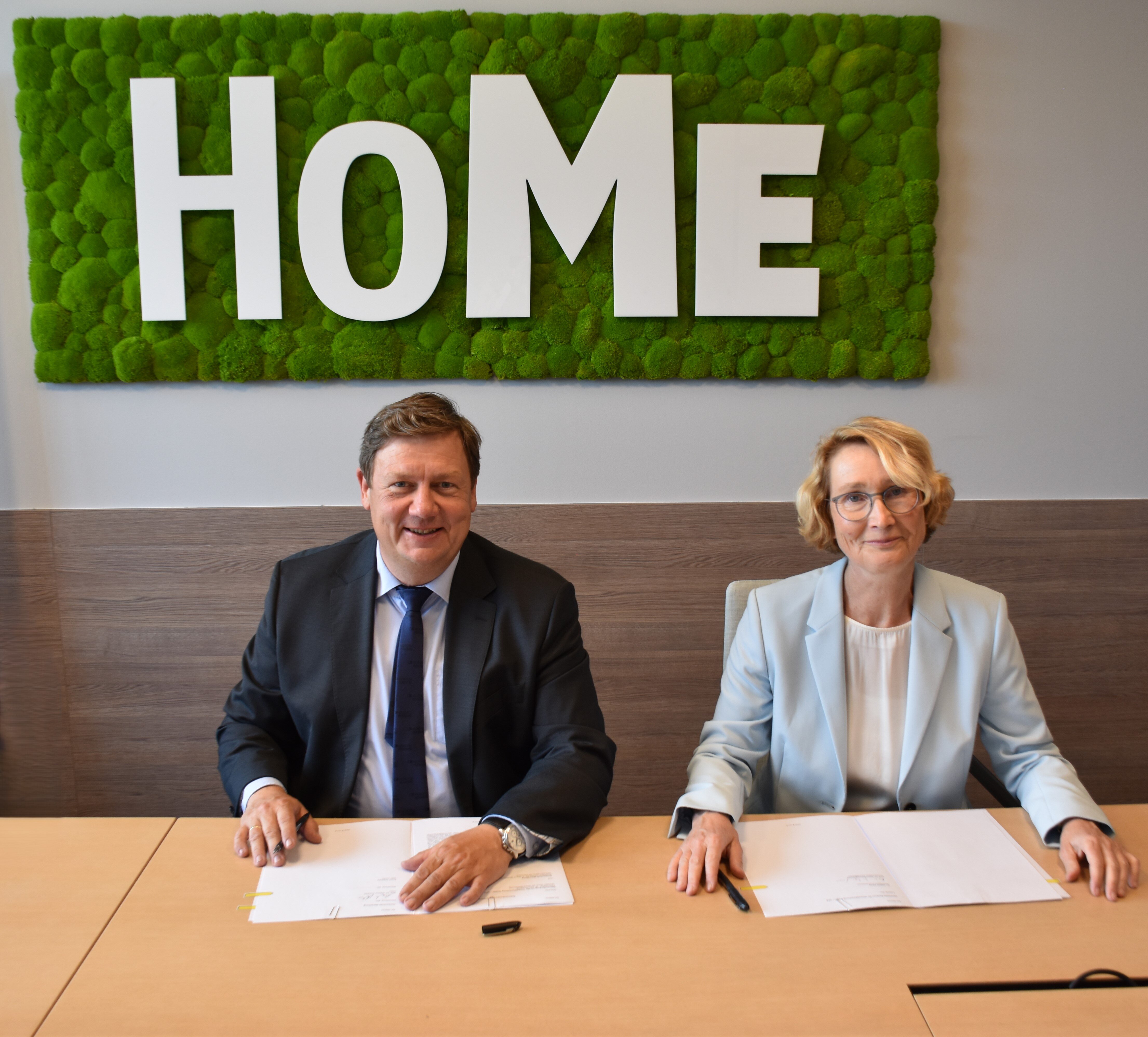 2 Personen an einem Tisch schauen in die Kamera. Im Hintergrund ein grüner Wandteppich mit der Aufschrift HoMe