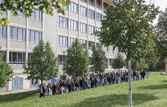 Eine große Gruppe von Studierenden laufen über den grünen Campus am Hauptgebäude der Hochschule Merseburg vorbei.