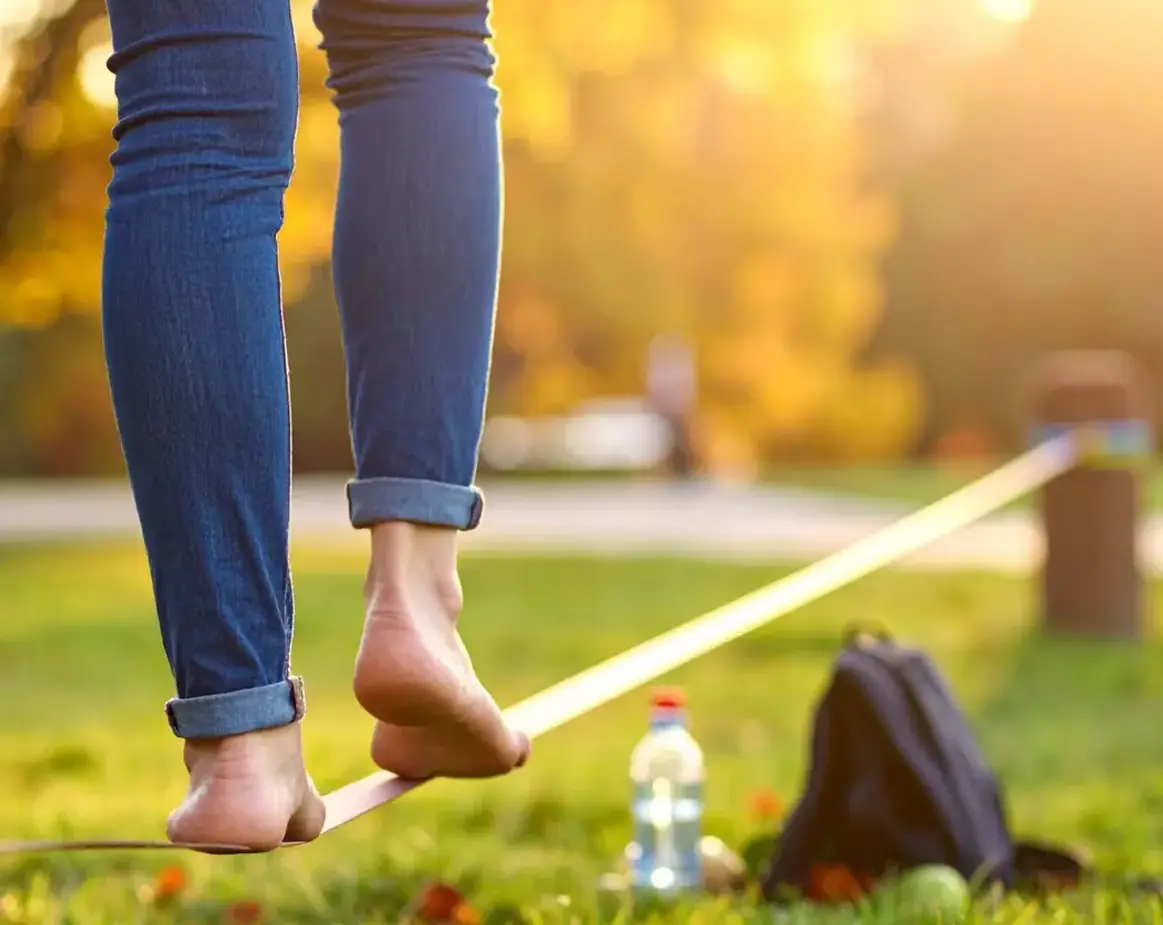 Eine Frau balanciert auf einer Slagline im Park. Man sieht nur die Beine als Ausschnitt von hinten.