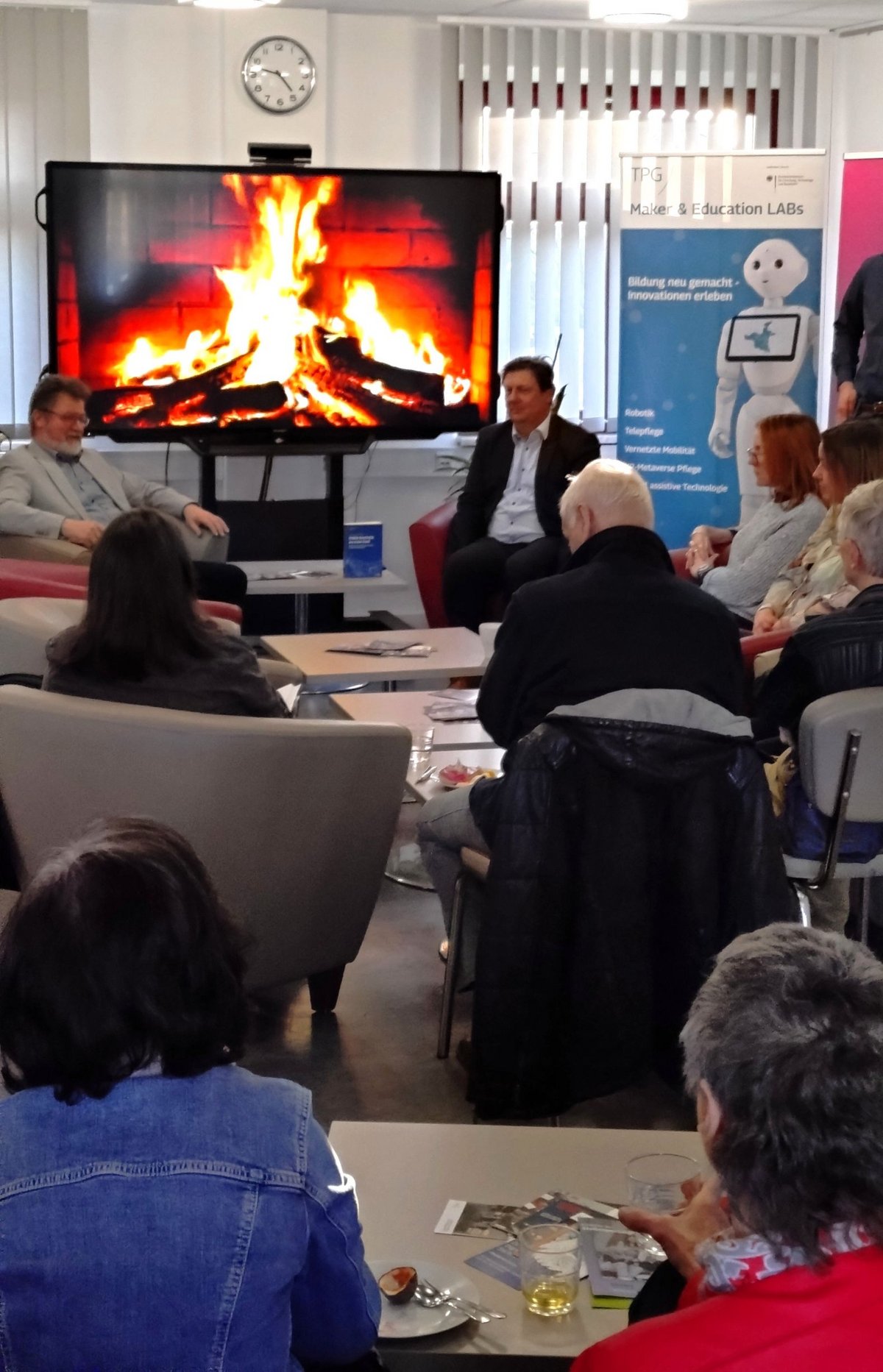 Blick von hinten auf das sitzende Publikum und dem Bildschirm mit dem digitalen Kamin