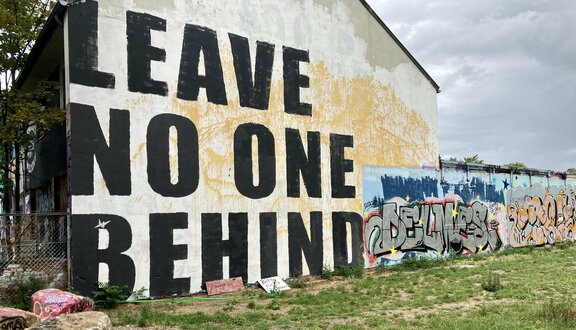 Eine Hausfassade, auf die der Schriftzug &quot;Leave No One Behind&quot; aufgemalt wurde. Rechts schließt sich eine Mauer mit bunten Graffitis an. 