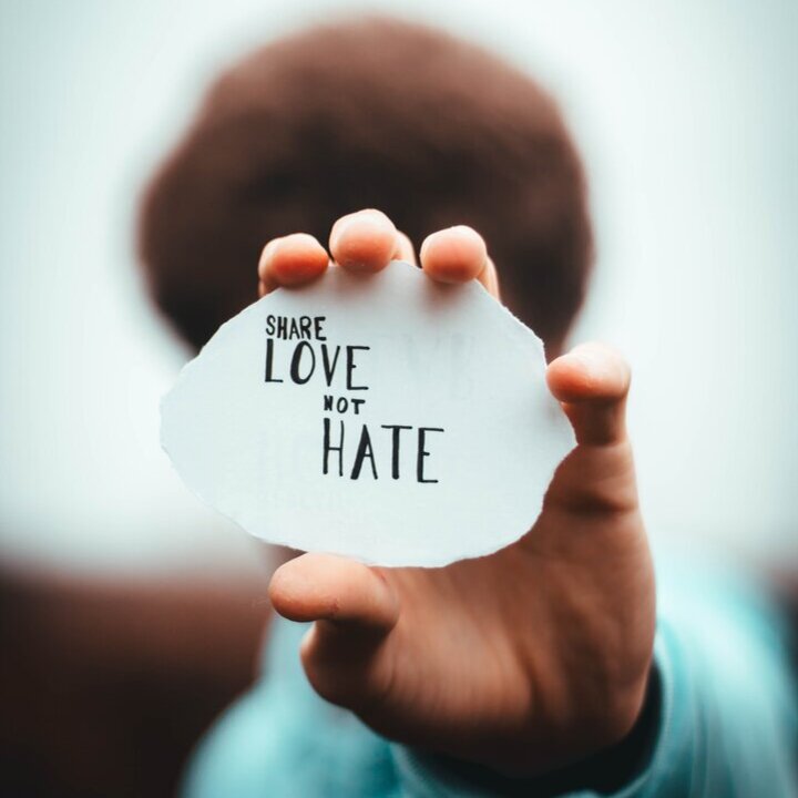 Person, die einen Zettel vor ihr Gesicht in die Kamera hält auf dem steht "Share love not hate"