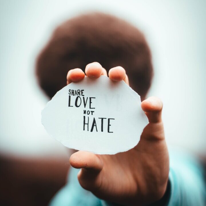 Person, die einen Zettel vor ihr Gesicht in die Kamera hält auf dem steht "Share love not hate"