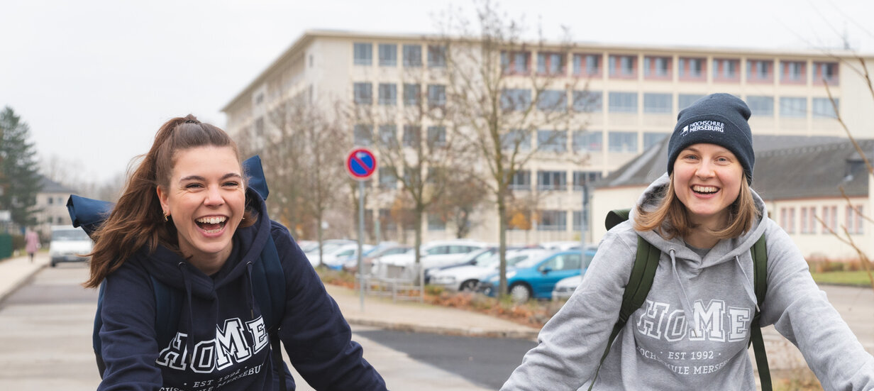 2 Personen lachend mit blauen und grauen Hochschulpullover an auf Fahrrädern im Vordergrund, im Hintergrund ein großes Gebäude mit Fenstern, Jahreszeit ist Herbst